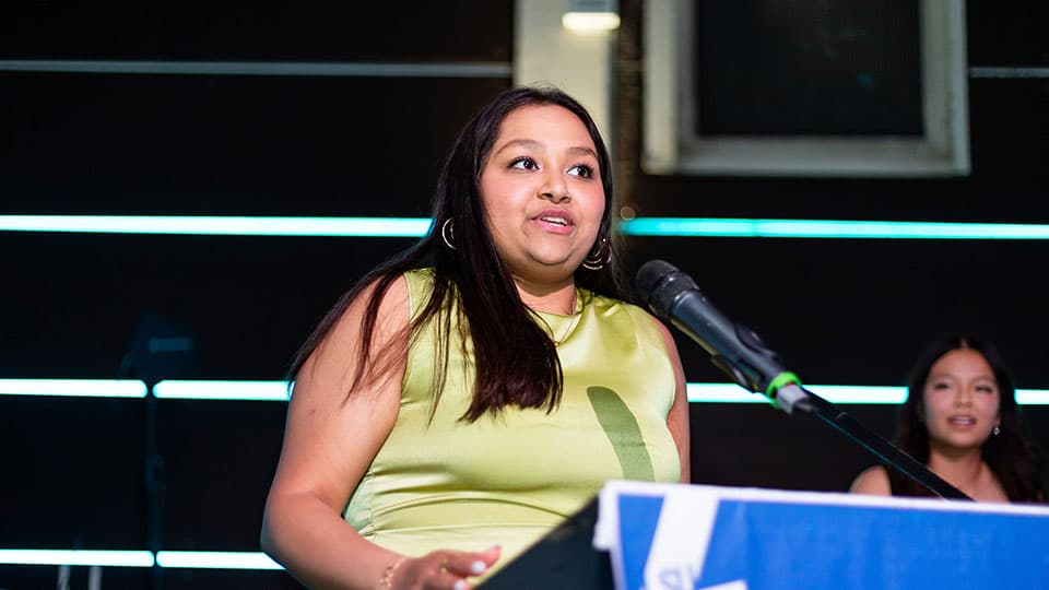 Tanishka is wearing a light green dress and is standing in front of a microphone delivering a speech on stage.